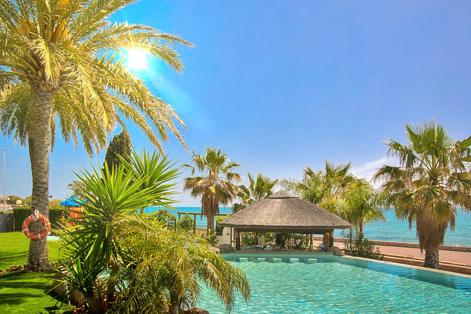 Vista amplia de piscina de arena con kiosco, palmeras y mar en un hotel