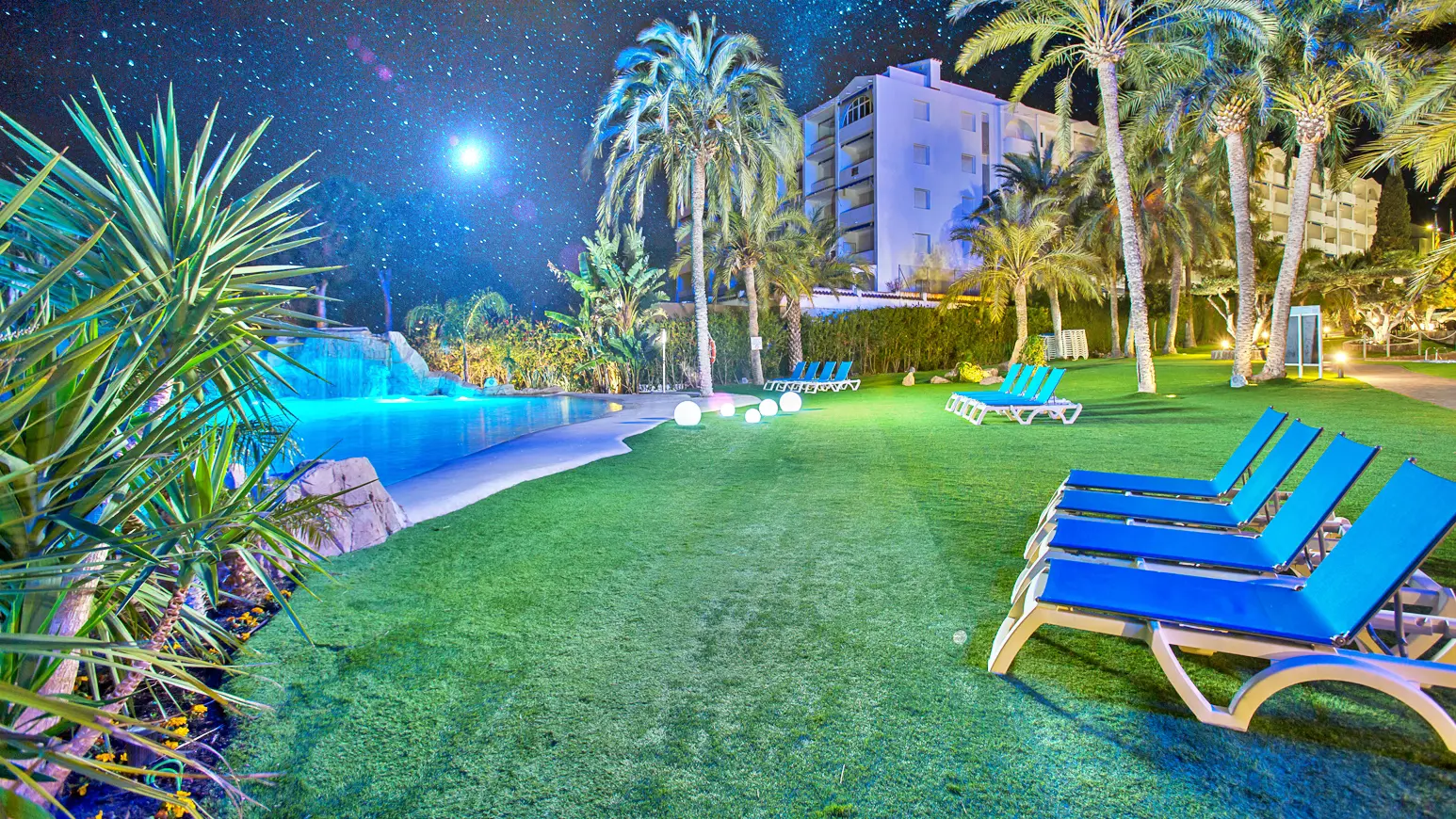 Piscina de arena iluminada de noche en un resort con palmeras y tumbonas
